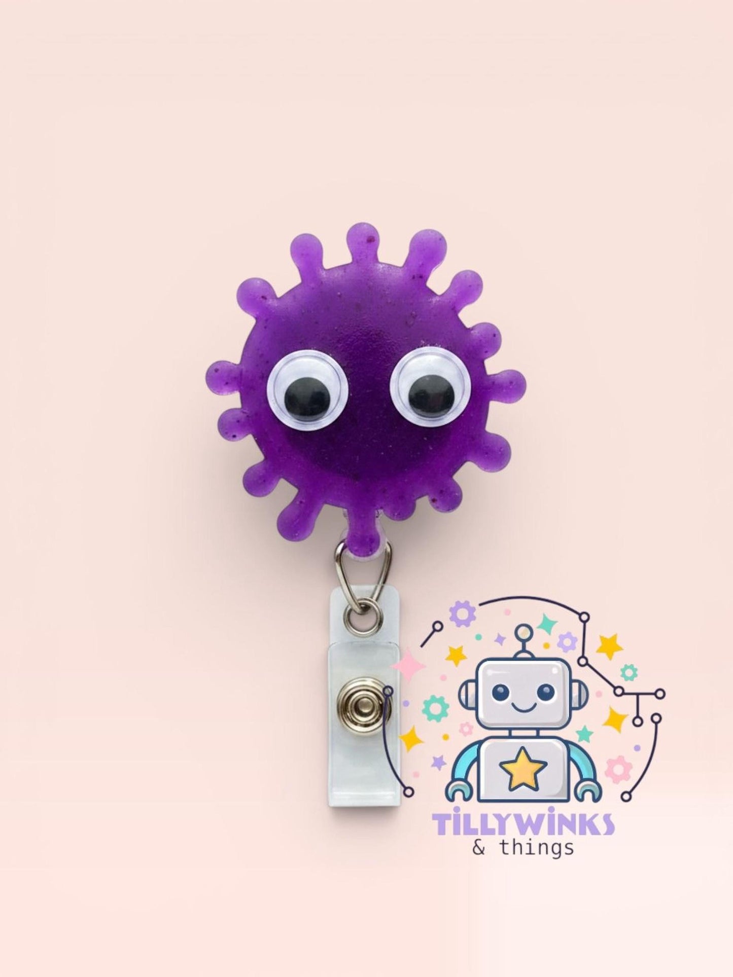a purple, googly-eyed character resembling a virus or bacteria, with a name tag attached to it. The character is hanging on a wall, and there is a logo or text in the background that reads "Tillywinks & things".