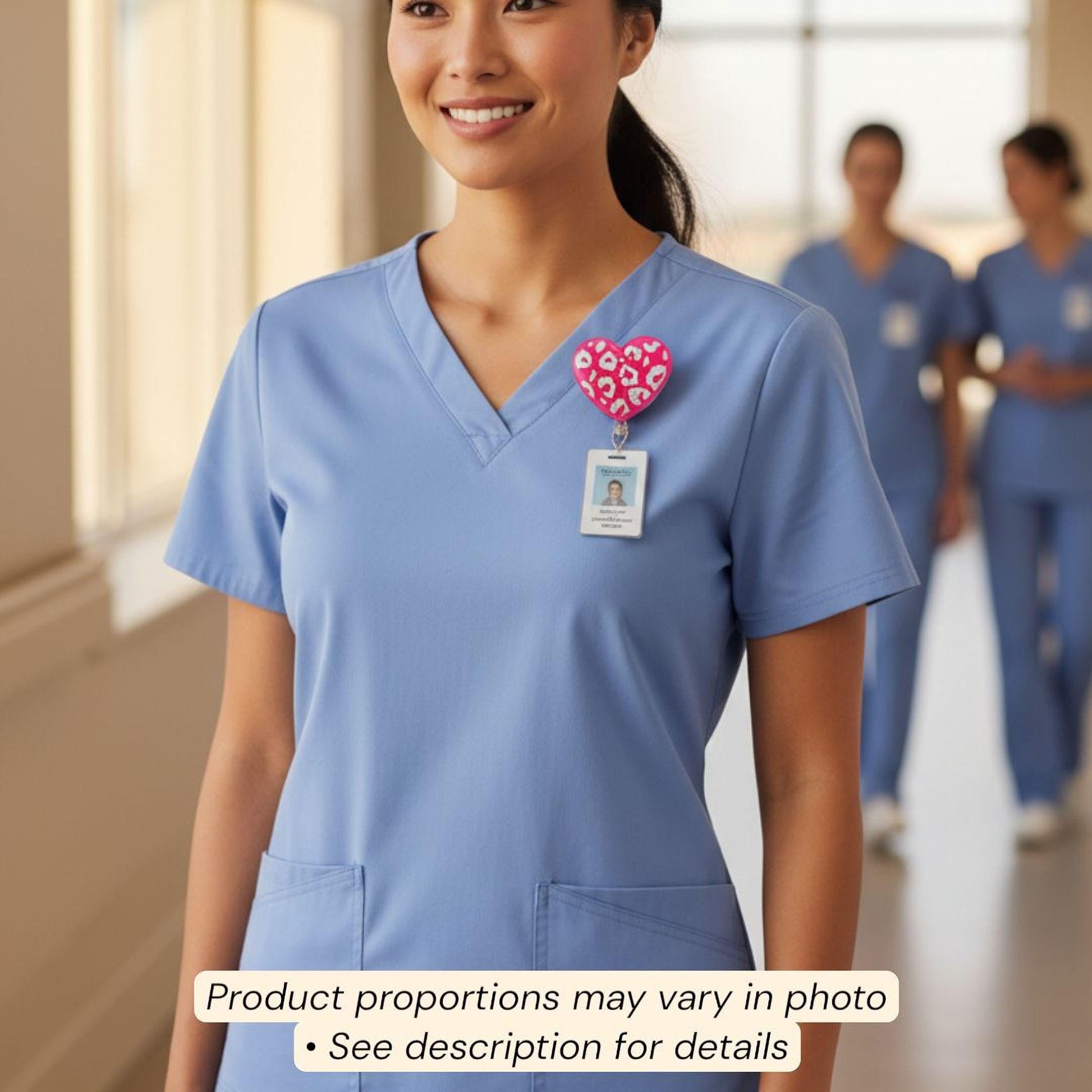 A smiling woman wearing blue scrubs with a heart-shaped badge on her shirt, standing in a hallway.
