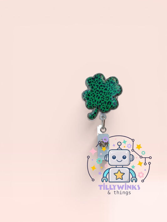 a green clover-shaped charm hanging from a metal clip, with a robot-like figure in the background.