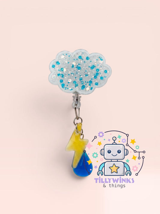 a cloud-shaped charm with blue and white glitter, hanging from a metal hook. Below the charm, there is a robot-like character with a yellow lightning bolt and a blue droplet, surrounded by various colorful elements.